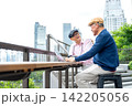 Two senior LGBTQ men engage in lively conversation at modern outdoor cafe with city skyline and lush greenery 142205056
