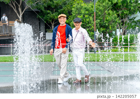Two senior LGBTQ men walk joyfully through cascading fountain water in vibrant urban park 142205057