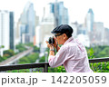 Senior LGBTQ man captures cityscape through vintage camera on modern balcony overlooking green park 142205059