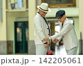 Two elderly men in white suits share a joyful moment, one adjusting the other's cuff with gentle hands. 142205061