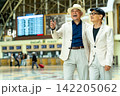 Two joyful senior men in matching white suits laugh together at train station, one pointing excitedly toward departure board. 142205062