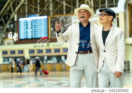 Two joyful senior men in matching white suits laugh together at train station, one pointing excitedly toward departure board. Two joyful senior men in matching white suits laugh together at train station, one pointing excitedly toward departure board. 142205062