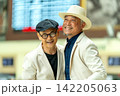 Two smiling senior men in stylish white jackets pose warmly at train station, one in hat, one in cap 142205063