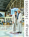 Two senior LGBTQ men in cream suits stand together at train station, one in hat smiling warmly 142205065