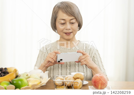リビングでスマホ操作しながら朝食のパンを食べるシニア女性 リビングでスマホ操作しながら朝食のパンを食べるシニア女性 142208259