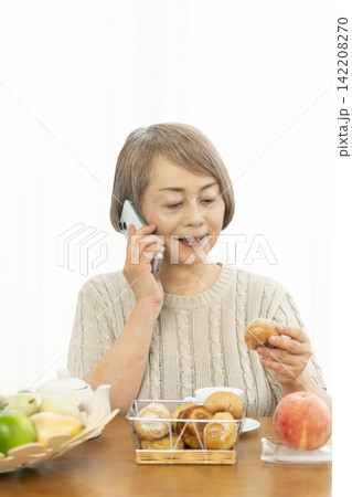 リビングでスマホ操作しながら朝食のパンを食べるシニア女性 142208270