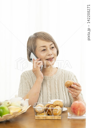 リビングでスマホ操作しながら朝食のパンを食べるシニア女性 142208271