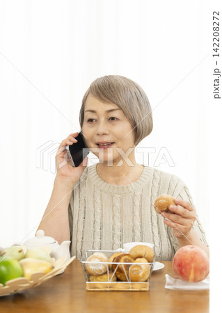 リビングでスマホ操作しながら朝食のパンを食べるシニア女性 142208272