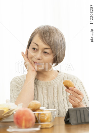 リビングでスマホ操作しながら朝食のパンを食べるシニア女性 142208287