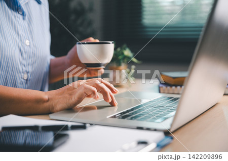 Person holding a coffee cup while using a laptop at a home workspace, representing remote work, online study, digital productivity, flexible employment, and modern work-life balance. Person holding a coffee cup while using a laptop at a home workspace, representing remote work, online study, digital productivity, flexible employment, and modern work-life balance. 142209896
