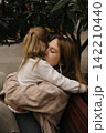 A mother hugs her little girl tightly while sitting on a bench among plants in a park. 142210440