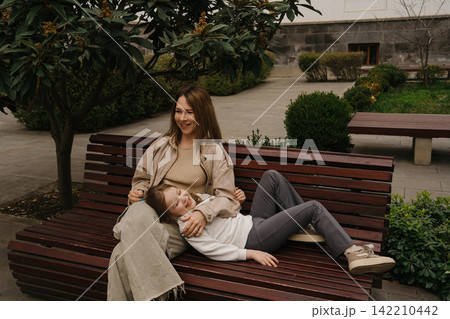Mom sits on a bench while daughter lays on her lap in a park during daytime 142210442