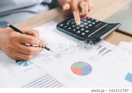 Business professional using a calculator while pointing at printed charts with a pen, representing financial analysis, accounting, budgeting, forecasting, performance review, 142212787