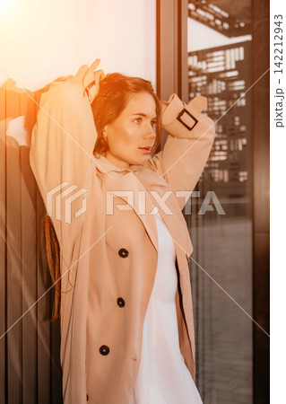 Woman fashion city posing in a beige trench coat against a glass building wall during sunset golden hour Woman fashion city posing in a beige trench coat against a glass building wall during sunset golden hour 142212943