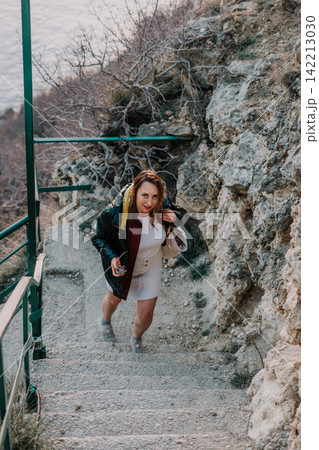 Woman climbing stairs on a rocky cliffside path near the coast while smiling at the camera 142213030
