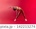 Yong woman in golden sportswear stretching her body over crimson backrground in studio 142213274