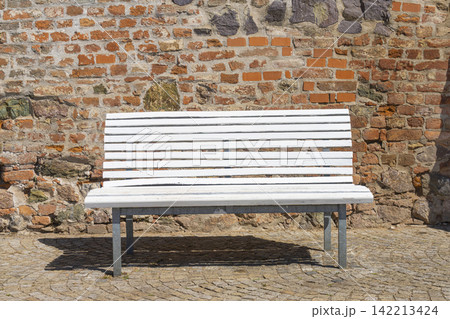 A white slatted park bench sits in front of a rough brick and stone wall. A white slatted park bench sits in front of a rough brick and stone wall. 142213424