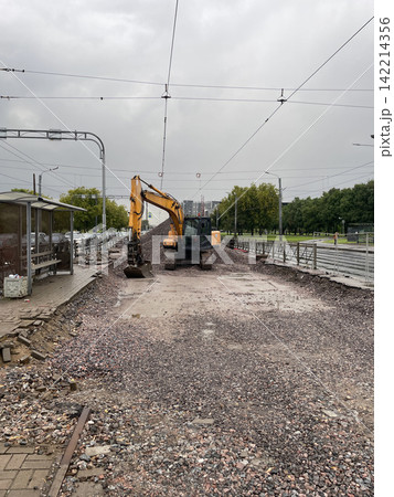 excavator working on construction site in urban area with tram tracks and trees in background. ground level, facing the excavator and construction site. industrial setting. excavator working on construction site in urban area with tram tracks and trees in background. ground level, facing the excavator and construction site. industrial setting. 142214356
