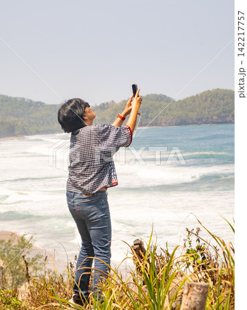 A female tourist crouching on the rocky shore and taking photos with his smartphone at Klayar Beach in Pacitan, East Java, Indonesia. A female tourist crouching on the rocky shore and taking photos with his smartphone at Klayar Beach in Pacitan, East Java, Indonesia. 142217757