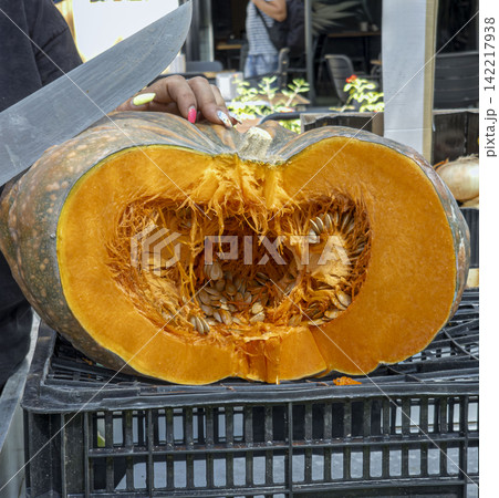 Chef holds large knife over halved pumpkin filled with seeds and pulp at outdoor market 142217938
