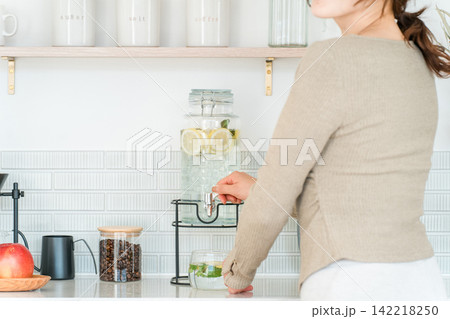 キッチンカウンターでウォーターサーバーからデトックスウォーターを注ぐ女性 キッチンカウンターでウォーターサーバーからデトックスウォーターを注ぐ女性 142218250