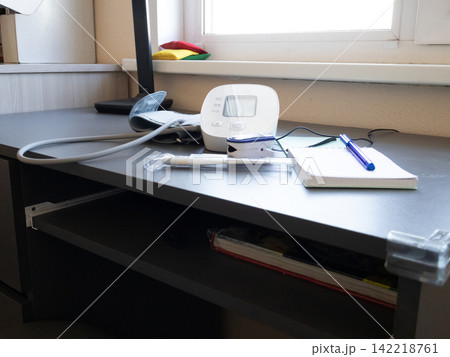Digital blood pressure monitor with cuff sits idle on a tidy desk by a window in daylight 142218761