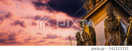 historic religious stone statues holding a golden cross and palm frond, silhouetted against a beautiful bright sunset sky with wispy white clouds in Prague. Detail of the Holy Trinity Column. Statue historic religious stone statues holding a golden cross and palm frond, silhouetted against a beautiful bright sunset sky with wispy white clouds in Prague. Detail of the Holy Trinity Column. Statue 142221551