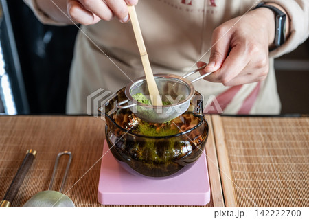 Close up of someone sift Matcha powder through a sifter into a bowl to remove any clumps. Matcha is a finely ground powder made from shade-grown green tea leaves. Close up of someone sift Matcha powder through a sifter into a bowl to remove any clumps. Matcha is a finely ground powder made from shade-grown green tea leaves. 142222700