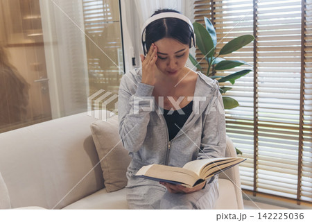Happy asian woman use headphones listen to music reading book sitting on cozy sofa in living room at home. Female asian people work at home research book read magazine smiling happy reader lifestyle Happy asian woman use headphones listen to music reading book sitting on cozy sofa in living room at home. Female asian people work at home research book read magazine smiling happy reader lifestyle 142225036