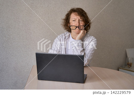 Woman Resting Hand Near Laptop While Looking Tired At Table In Plain Indoor Room With Beige Wall Woman Resting Hand Near Laptop While Looking Tired At Table In Plain Indoor Room With Beige Wall 142229579
