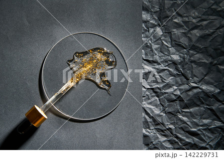 Black Handled Dropper Laying Across Round Glass Dish With Gold Flake Gel On Split Textured Black Black Handled Dropper Laying Across Round Glass Dish With Gold Flake Gel On Split Textured Black 142229731