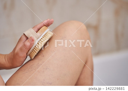 Woman Holding Wooden Bristle Brush On Leg In Bathtub Demonstrating Circular Motion For Dry Brushing Woman Holding Wooden Bristle Brush On Leg In Bathtub Demonstrating Circular Motion For Dry Brushing 142229881