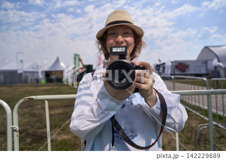 Smiling Woman Holding Camera At Outdoor Fairground Fence Under Bright Cloudy Sky During Daytime Smiling Woman Holding Camera At Outdoor Fairground Fence Under Bright Cloudy Sky During Daytime 142229889
