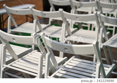 White Folding Chairs Clustered Together In Sunlit Outdoor Seating Area Before Ceremony On Warm Day White Folding Chairs Clustered Together In Sunlit Outdoor Seating Area Before Ceremony On Warm Day 142229890