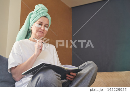 Woman Reviewing Journal In Bedroom Wearing Towel Turban And Eye Patches While Seated Crosslegged Woman Reviewing Journal In Bedroom Wearing Towel Turban And Eye Patches While Seated Crosslegged 142229925