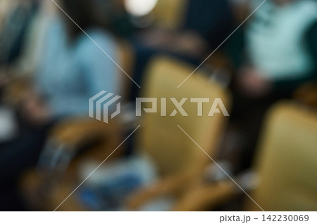 Blurred Audience Sitting In Conference Chairs Inside Indoor Seminar With Warm Ambient Lighting Blurred Audience Sitting In Conference Chairs Inside Indoor Seminar With Warm Ambient Lighting 142230069