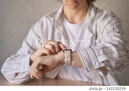 Adult Woman Checking Wristwatch At Desk In Plain Indoor Office Setting, Wearing Striped Shirt Adult Woman Checking Wristwatch At Desk In Plain Indoor Office Setting, Wearing Striped Shirt 142230209