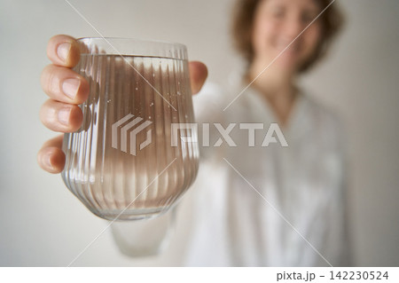 Closeup Glass Of Water Held In Foreground By Smiling Woman In Soft Indoor Light With Blurred Closeup Glass Of Water Held In Foreground By Smiling Woman In Soft Indoor Light With Blurred 142230524