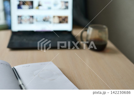 Notebook With Pen And Coffee Stain Beside Blurred Laptop Screen And Glass Mug On Wooden Desk 142230686