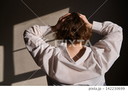 Young Woman Holding Head With Raised Elbows In Sunlit Interior Beside Window Shadow On Neutral Young Woman Holding Head With Raised Elbows In Sunlit Interior Beside Window Shadow On Neutral 142230699