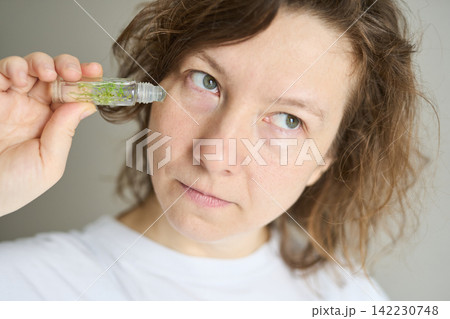 Woman Applying Rollon Under Eye In Neutral Indoor Setting Holding Small Glass Vial With Green Herbs Woman Applying Rollon Under Eye In Neutral Indoor Setting Holding Small Glass Vial With Green Herbs 142230748