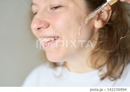 Woman Applying Clear Facial Serum With Pipette To Lower Cheek While Smiling And Looking Slightly 142230994