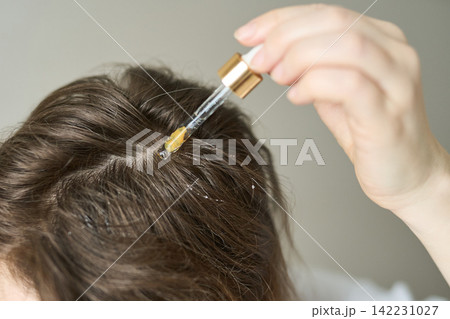 Woman Applying Clear Scalp Serum With Glass Dropper To Parted Brown Hair Roots In Closeup Indoor Woman Applying Clear Scalp Serum With Glass Dropper To Parted Brown Hair Roots In Closeup Indoor 142231027