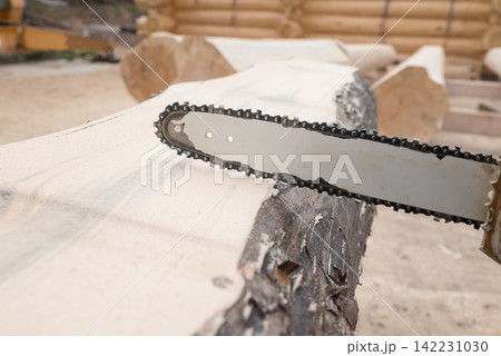 A professional lumberjack saws logs, holding a chainsaw in his hand, to build a wooden house. The construction site of a wooden house. A professional lumberjack saws logs, holding a chainsaw in his hand, to build a wooden house. The construction site of a wooden house. 142231030