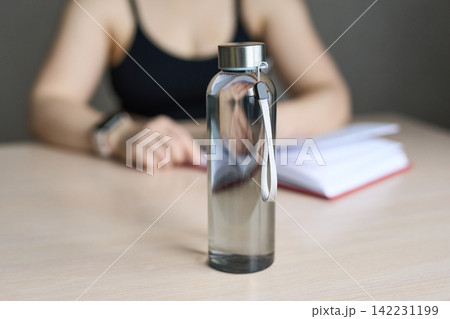 Tall Clear Water Bottle Standing On Wooden Table With Blurred Person In Sleeveless Top Writing Tall Clear Water Bottle Standing On Wooden Table With Blurred Person In Sleeveless Top Writing 142231199