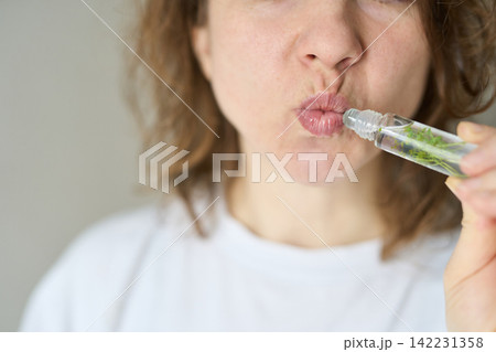 Woman Puckering Lips Applying Roller Lip Gloss With Botanical Fragments In Transparent Bottle 142231358
