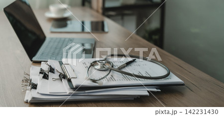 Stethoscope rests on printed documents beside laptop and pen on wooden desk, with clipboard and paper stack arranged for medical documentation, healthcare records, and clinical management. Stethoscope rests on printed documents beside laptop and pen on wooden desk, with clipboard and paper stack arranged for medical documentation, healthcare records, and clinical management. 142231430
