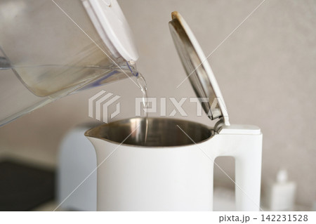 White Kettle Being Filled With Water From Plastic Pitcher On Kitchen Countertop With Open Lid White Kettle Being Filled With Water From Plastic Pitcher On Kitchen Countertop With Open Lid 142231528