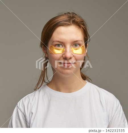 Young Woman Wearing Gold Eye Patches Facing Camera With Gentle Smile And Relaxed Expression Young Woman Wearing Gold Eye Patches Facing Camera With Gentle Smile And Relaxed Expression 142231535