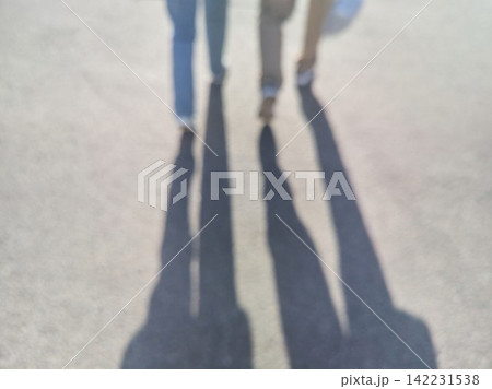 Two People Casting Long Blurred Shadows While Walking On Sunlit Pavement Producing Elongated Two People Casting Long Blurred Shadows While Walking On Sunlit Pavement Producing Elongated 142231538